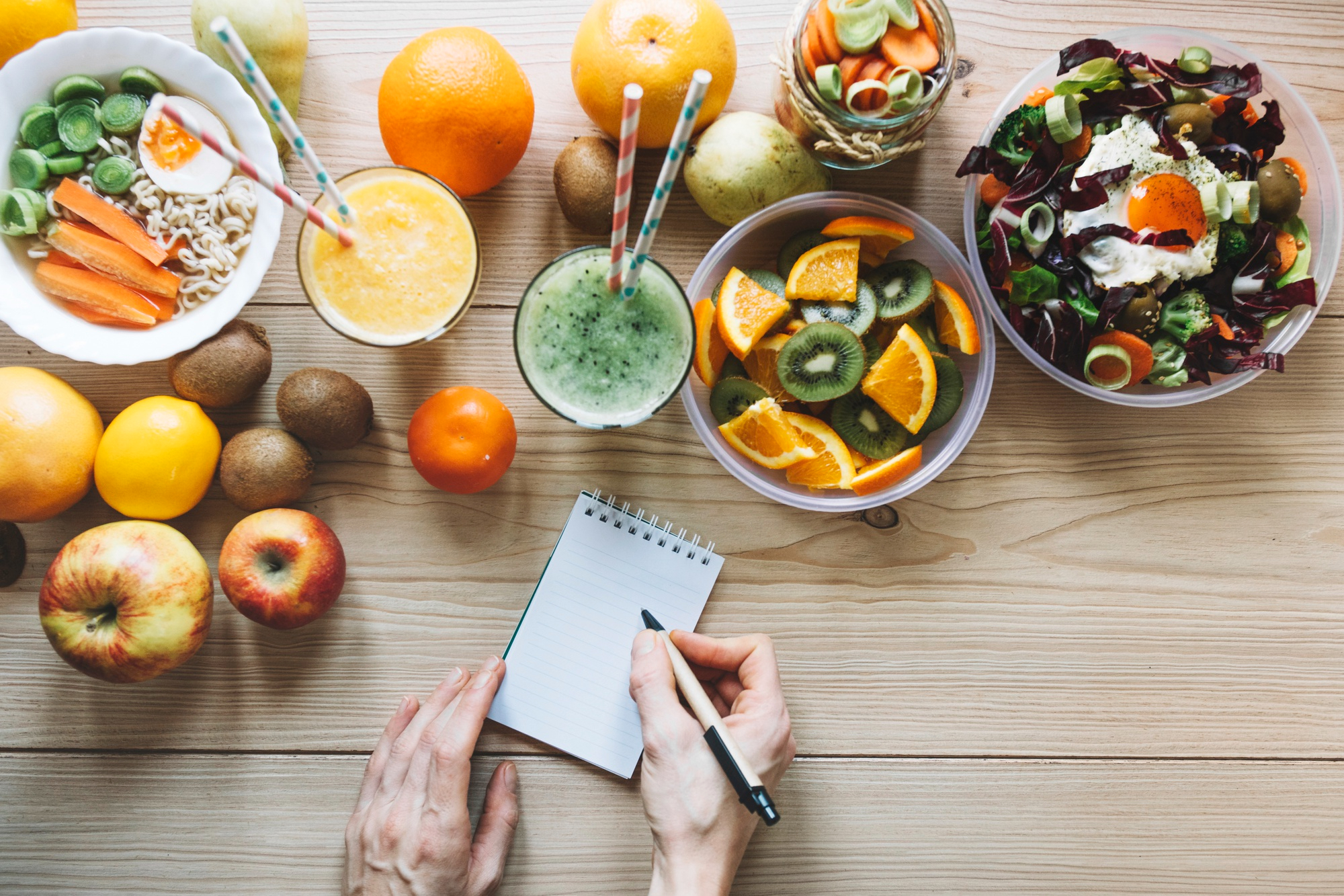 Mesada con alimentos saludables, frutas, verduras, licuados y jugos naturales. Manos de una persona tomando nota sobre consejos de salud.
